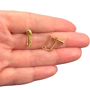 May include: Two gold-coloured clip-on earring findings. One is a simple clip, and the other has a screw-back mechanism. The findings are designed for creating clip-on earrings. The background is a person's hand.