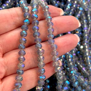 Op de afbeelding: Close-up van strengen iriserende, gefacetteerde kralen in grijze, paarse en blauwe tinten. De kralen worden in een hand gehouden, waardoor hun reflecterende aard en ingewikkelde snit worden benadrukt. Deze kralen zijn waarschijnlijk bedoeld voor het maken van sieraden of knutselen.