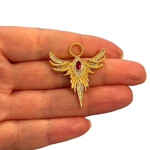 May include: A gold-coloured angel brooch featuring a central oval ruby-coloured stone and small clear crystals. The angel has outstretched wings and a halo. The brooch is held in a hand against a white background.