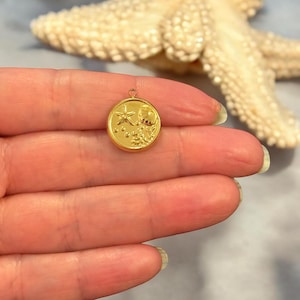 May include: A small, round gold charm with a starfish, bubbles, and other sea life designs. The charm has a loop at the top for attaching to a necklace or bracelet. The charm is held in a person's hand.