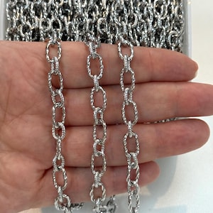 May include: A silver-toned metal chain featuring elongated oval links with a distinct twisted, textured surface pattern. The chain is composed of repeating, uniform links that create a continuous, decorative strand suitable for jewelry making.