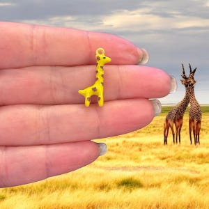 Puede incluir: Un pequeño amuleto de jirafa amarillo con manchas marrones. El amuleto está siendo sostenido en una mano, con un fondo de sabana con dos jirafas en la distancia.