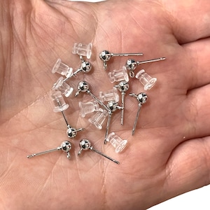 May include: A selection of silver stud earrings with clear plastic backs. Each earring has a small, round, silver ball at the end of a thin post. The earrings are arranged on a light surface, possibly for jewellery making or craft purposes.