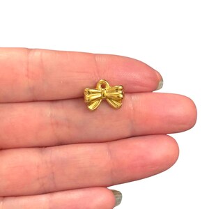 Pode incluir: Um pequeno berloque de laço dourado com uma argola na parte superior para prender a joias. O laço tem um design detalhado e texturizado e é mostrado em um fundo branco.
