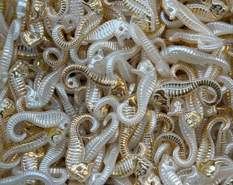May include: A close-up shot of a pile of seahorse-shaped beads. The beads are a mix of white and gold, with the gold accents highlighting the details of the seahorse design. The beads are small and appear to be made of plastic or resin.