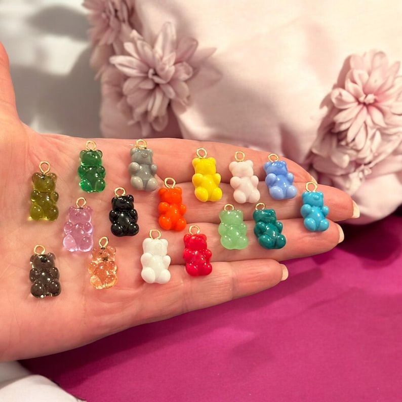 May include: A collection of colorful gummy bear charms in various colors, including green, yellow, and blue. Each charm has a gold-colored loop at the top. The charms are held in a hand against a soft pink background.