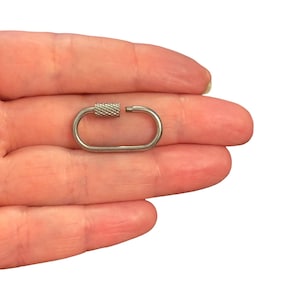 May include: A small, oval-shaped silver-toned metal carabiner with a threaded screw-lock closure. The locking mechanism features a textured, cross-hatched grip pattern, and the gate is shown in an open position, revealing the small threaded end.