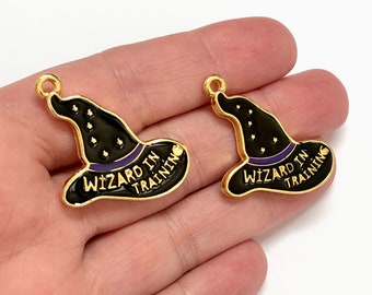 Op de afbeelding: Twee gouden metalen bedels in de vorm van zwarte heksenhoeden met paarse bies. Elke bedel heeft de tekst "Wizard in Training" op de rand.