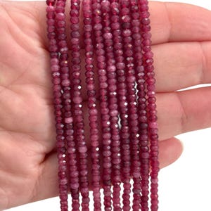 May include: Multiple strands of faceted, deep red gemstone beads. The small, uniform beads are strung together, creating a flowing, textured appearance. The colour is a rich, saturated red, with slight tonal variations. The beads are held in a hand.