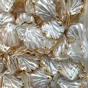 Puede incluir: Primer plano de un montón de cuentas blancas nacaradas en forma de concha con ribete dorado. Cada cuenta tiene un agujero en la parte superior para ensartar. Las cuentas tienen un acabado brillante y son ideales para la fabricación de joyas y manualidades.