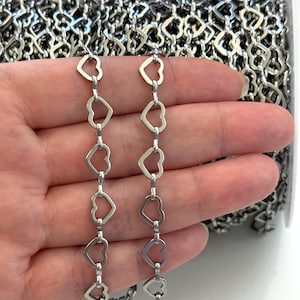 Puede incluir: Una cadena de metal en tono plateado compuesta por eslabones repetitivos en forma de corazón abierto. La cadena se presenta como una bobina continua, con un acabado pulido que refleja la luz en cada eslabón de corazón del diseño.