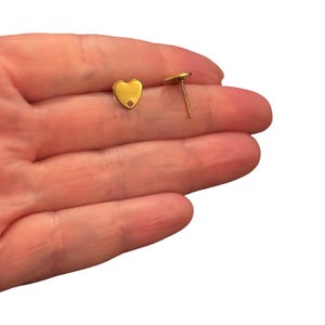 Könnte beinhalten: Zwei goldfarbene Ohrstecker in Herzform. Ein Ohrstecker zeigt die Vorderseite mit einem kleinen kreisförmigen Loch an der unteren Spitze, während der andere seitlich positioniert ist, um den dünnen Metallstift auf der Rückseite zu zeigen.