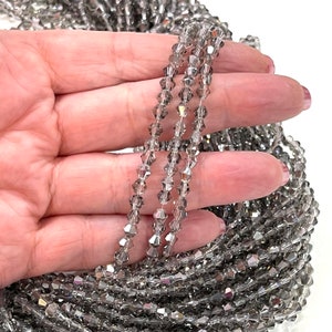 May include: A strand of silver crystal beads, with a faceted, teardrop shape. The beads are strung on a clear thread and are shown in a hand.