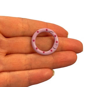 May include: A light purple metal clasp with a circular design. The clasp is decorated with small, evenly spaced pink dots. The clasp is held in a hand.