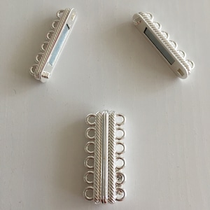 Puede incluir: Tres cierres magnéticos plateados para joyería. Dos cierres son rectangulares con una fila de bucles en un lado y una barra magnética. El tercer cierre es un cierre rectangular más grande con múltiples bucles y un cierre magnético.