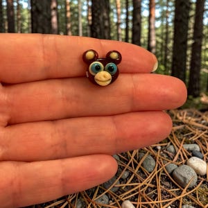 May include: A small, dark brown glass bead shaped like a bear head with large, bright blue eyes, a cream-colored snout, and two rounded ears with cream-colored centers. The bead features a smooth, glossy finish and a cartoon-like facial expression.