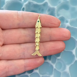 Peut inclure: Un pendentif en forme de poisson de couleur or avec un œil en pierre verte. Le pendentif a un design en couches, avec une petite boucle en haut pour une chaîne. La queue du poisson est en forme d'éventail fendu.