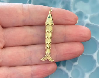 May include: A gold-colored fish pendant with a green gem eye. The pendant has a layered design, with a small loop at the top for a chain. The fish's tail is a split, fan-like shape.
