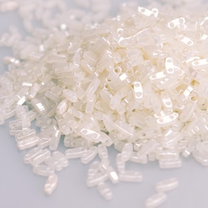 May include: A large pile of white seed beads, also known as bugle beads, with a slightly iridescent finish. The beads are small and cylindrical with a hole at each end.