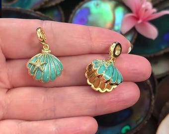 May include: Two gold-colored seashell charms with turquoise enamel and a starfish accent. One charm has a single shell, and the other has a layered shell design. The charms are displayed against a blurred background.