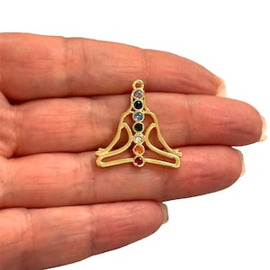 May include: A gold-coloured pendant in the shape of a person in a seated yoga pose. The pendant features seven colourful gemstones aligned vertically, representing the chakras. The gemstones are purple, dark blue, light blue, green, white, orange, and red.