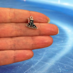 May include: A miniature silver charm depicting a black ice skate with red detailing and a star design, mounted on a silver runner. The charm has a loop for attachment. The charm is held in a hand. The charm is a winter-themed accessory.