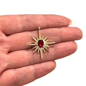 May include: A gold-coloured pendant in the shape of a starburst, with a central oval red gemstone. The pendant has multiple pointed rays extending outwards, forming a star. The pendant is held in a hand.