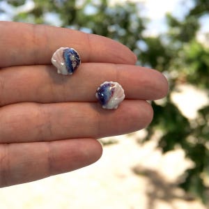 Pode incluir: Dois brincos pequenos em forma de concha. Os brincos são brancos com detalhes em rosa, azul e roxo. São segurados em uma mão, com um fundo verde desfocado.