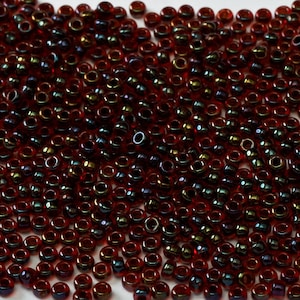 May include: A large quantity of small, round, iridescent red beads. The beads have a metallic sheen and are scattered across a white surface.