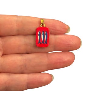 May include: A rectangular pendant featuring a vibrant red enamel background with three stylized blue fish arranged vertically. The pendant is framed in gold-tone metal and includes a small gold-tone jump ring at the top for attachment to a chain.