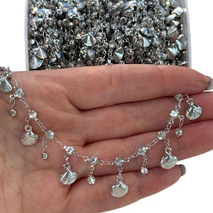 Könnte beinhalten: Silberkette mit klaren Kristallperlen und silbernen Muschel-Anhängern. Die Kette wird auf einer Hand gezeigt.