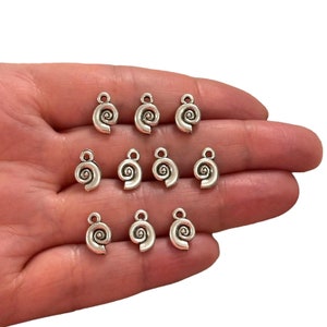Puede incluir: Doce abalorios de metal plateados en forma de conchas con un diseño en espiral. Los abalorios tienen un bucle en la parte superior para sujetarlos a joyas u otras manualidades.