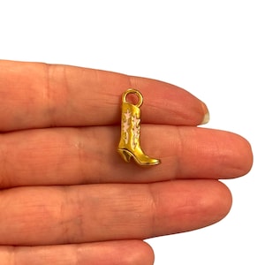 May include: A small, gold-colored cowboy boot charm with pink floral accents. The charm has a loop at the top for attaching to a necklace or bracelet. The boot has a pointed toe and a small heel.