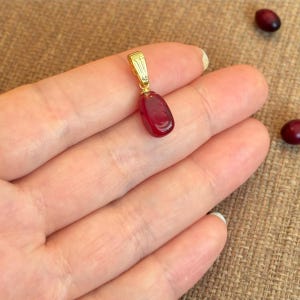 May include: A gold-colored pendant with a teardrop-shaped, deep red gemstone. The pendant is suspended from a small, rectangular bail. The gemstone is smooth and polished, reflecting light. The background is a neutral, textured surface.