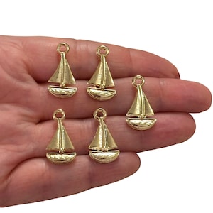 Op de afbeelding: Zes gouden zeilbootbedels met een gestructureerd oppervlak. De bedels zijn klein en hebben een lus aan de bovenkant om aan sieraden of andere knutselwerkjes te bevestigen.