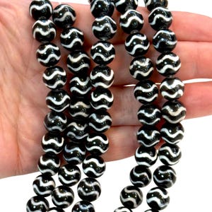 May include: Close-up of black and white patterned beads. Each bead is round with a glossy finish and a swirling white design. The beads are strung together, possibly for a necklace or bracelet. The background is a neutral colour.