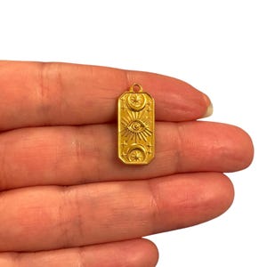 May include: A gold-colored pendant with an eye, sun rays, crescent moons, and stars design. The rectangular pendant has a small loop at the top for a chain. The pendant is held in a hand against a white background.