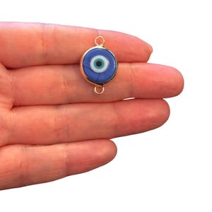 May include: A circular evil eye charm featuring a blue, white, and black concentric design encased in a gold-toned metal frame. The charm includes two small gold-toned loops, one at the top and one at the bottom, for attachment purposes.