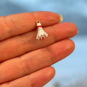 May include: A miniature white and red badminton shuttlecock charm with a gold-colored loop. The shuttlecock has a white feathered skirt and a red band near the top. The charm is designed for jewelry making or crafting.