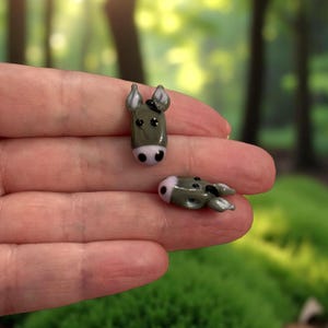 Może przedstawiać: Dwie miniaturowe szklane głowy osłów. Jedna jest w pozycji pionowej, druga leży. Osły są szare z różowymi pyskami i czarnymi uszami. Tło jest rozmyte zielone.