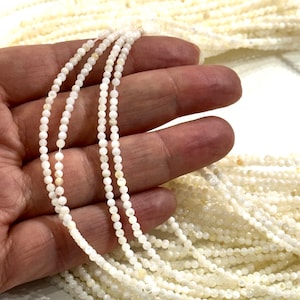 Può includere: Primo piano di più fili di piccole perline rotonde bianco sporco. Le perline sono infilate insieme, creando un aspetto delicato ed elegante. Le perline sono tenute in una mano, con più fili visibili sullo sfondo. Le perline sono di colore bianco crema.