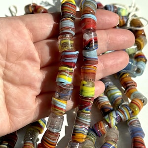 Può includere: Due fili di perline cilindriche in vetro con un motivo marmorizzato dai colori vorticosi rosso, giallo, blu, marrone e bianco. Le perline hanno una finitura lucida e traslucida e sono infilate su un filo bianco, mostrando una varietà di bande di colore orizzontali.