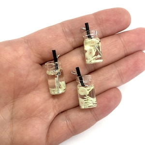 Peut inclure: Trois petits pots en verre avec un liquide clair et des tranches de fleurs blanches à l'intérieur. Chaque pot a une paille noire et un couvercle argenté. Les pots sont sur une main.