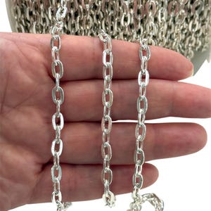 Può includere: Primo piano di una catena color argento con maglie ovali. La catena è tenuta in mano, con un rotolo più grande della stessa catena visibile sullo sfondo. La catena viene probabilmente utilizzata per la creazione di gioielli o altri lavori artigianali.