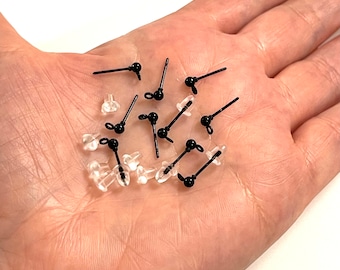 May include: A close-up of a hand holding a group of black earring backs. The backs are made of plastic and have a small metal loop on the top. The backs are designed to be used with stud earrings.