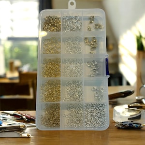 May include: A clear plastic storage box filled with jewellery-making components. The compartments contain gold and silver-coloured jump rings, clasps, and small beads. The box is on a wooden surface, with other craft tools visible in the background.