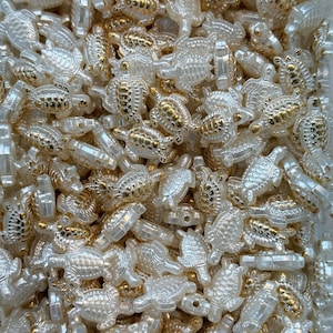 May include: A collection of small, translucent turtle-shaped beads with gold-colored accents. The beads are scattered, creating a textured, shimmering effect. These decorative beads are ideal for jewelry making and craft projects.