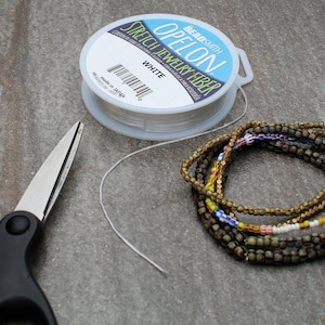 Könnte beinhalten: Eine Spule weißen "OPELON" Stretch-Schmuckfasers, eine Schere und Perlenarmbänder. Die Armbänder haben goldene, grüne und blaue Perlen. Die Schere ist silber und schwarz.