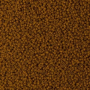 May include: A dense collection of small, uniform, cylindrical seed beads in a solid, matte mustard-brown color. Each bead features a central hole, creating a consistent, textured surface pattern across the entire frame of the image.