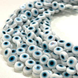 May include: A close-up shot of a collection of white beads, each featuring a blue and black eye design. The beads are round and have a glossy finish. They are strung together, forming a flowing pattern. These beads are likely used for jewellery making or other crafts.